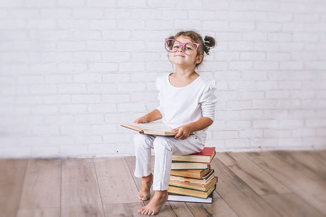 A child wearing glasses holds a book and sits on a stack of books