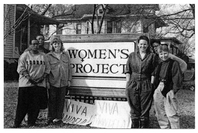 Six women, three on each side, stand next to an outdoor sign that reads, "Women's Project" while white posters rest on the ground below and read "Viva Mandela!"