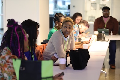 A young person leans forward and speaks to two other people amid a large group of people sitting around conference tables placed in a square conference configuration.