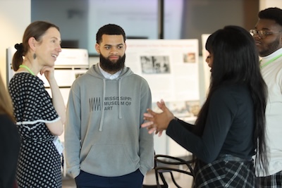 A young person stands in front of a poster presentation and listens as another person talks and gestures with their hands. Other posters and presenters appear in the background.